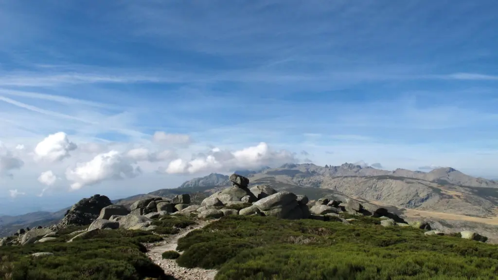 Sendero sobre la cresta de un lomo hacia formaciones rocosas, con una impresionante vista de la Sierra de Gredos al fondo