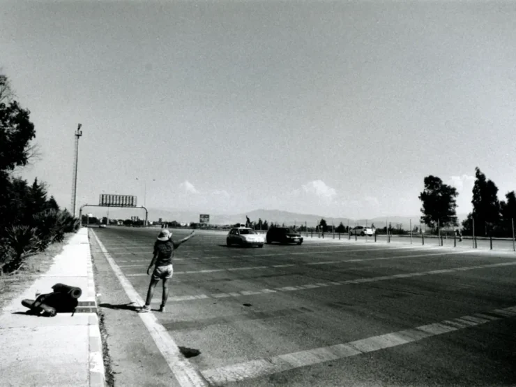 Foto en blanco y negro de un autoestopista junto a una gran carretera en un paisaje llano