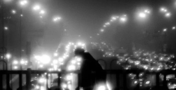 Foto blanco y negro de un paisaje urbano nocturno: un individuo con la cabeza doblada cruza un puente de la circunvalación