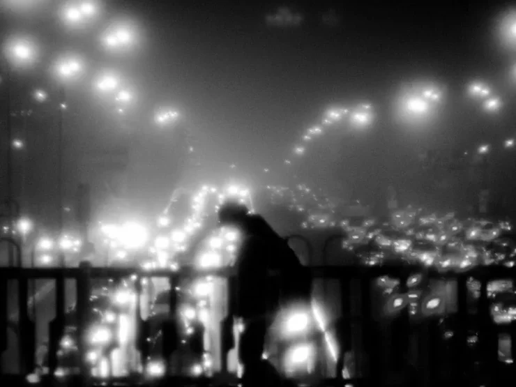 Foto blanco y negro de un paisaje urbano nocturno: un individuo con la cabeza doblada cruza un puente de la circunvalación