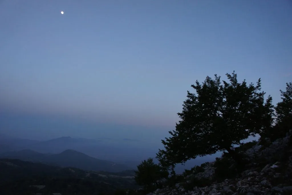 Atardecer brumoso, montañas cubiertas de hayas y una luna creciente alta en el cielo.