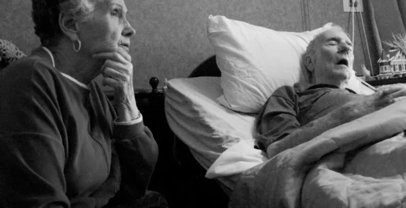 Pareja de mayores. La esposa sienta pensativa, el marido enfermo duerme en una cama