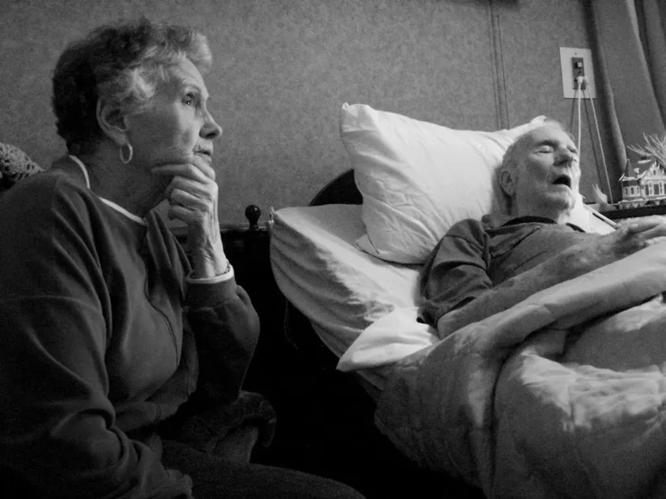 Pareja de mayores. La esposa sienta pensativa, el marido enfermo duerme en una cama
