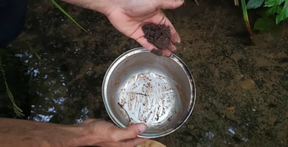 Mano sujetando una olla y una cuchara de madera sucias antes de fregarla sin jabón, con un poco de barro y agua de río