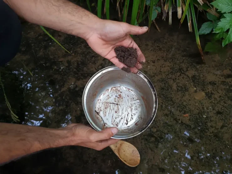 Mano sujetando una olla y una cuchara de madera sucias antes de fregarla sin jabón, con un poco de barro y agua de río