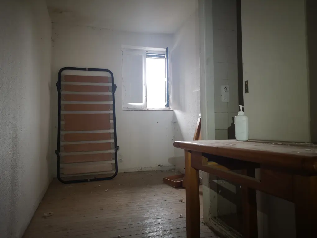 Habitación del albergue con el somier de una cama apoyada en la pared, una mesa de madera y una ventana abierta que entra mucha luz.