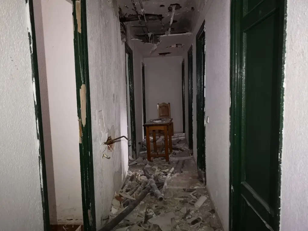 Pasillo en el albergue, los muros se han derrumbado y la cal cuelga del techo. Al fondo hay una silla y una mesa.
