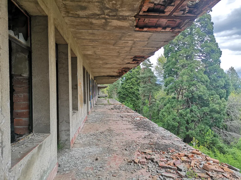 Enorme balcón sin barandilla en la fachada trasera del sanatorio abandonado de Tablada, con escombros, ventanas sin marcos y bosque de pinos a la izquierda