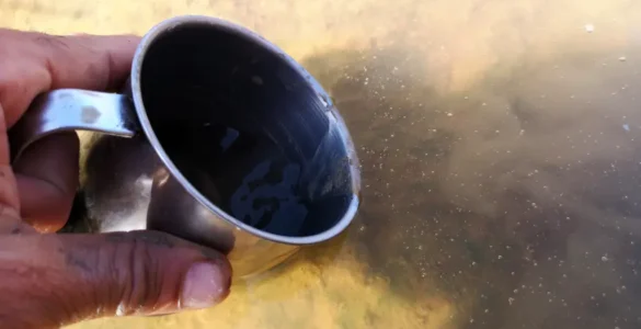 Mano recoge agua turbia del arroyo Pedraza con una taza de metal antes del proceso de filtrado y potabilización