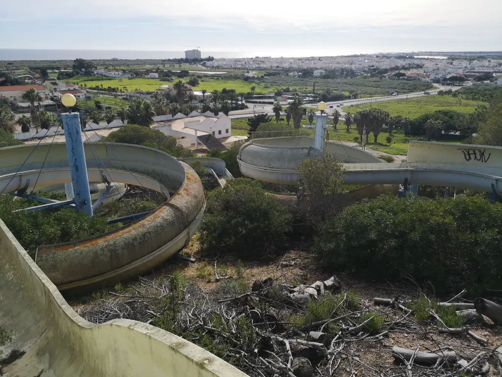 Vista aérea de los toboganes del parque acuático abandonado en Castro Marín, Portugal, con estructuras deterioradas y sin mantenimiento, rodeadas de vegetación, y el mar visible al fondo en el horizonte.
